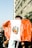 A person is wearing a large flag over their shoulders that features a coat of arms, possibly from Peru. The setting appears to be a busy urban street with a tall building and several people visible in the background. Light reflects off the flag, indicating it's a bright day.