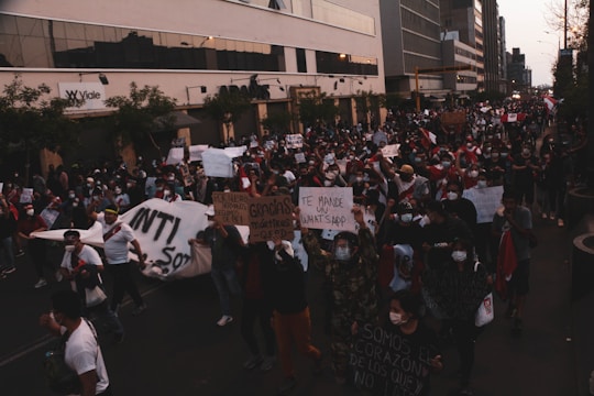 A large group of people is gathered in a protest or demonstration on a city street. They are holding signs and banners, some of which are in Spanish. Many participants are wearing face masks, and several are dressed in red and white clothing. The setting is urban, with tall buildings lining the street.