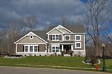 A two-story house is set against a backdrop of bare trees under a cloudy sky. The house features taupe siding with white trim and has wreaths hanging on the door and windows, suggesting a festive season. The front yard is well-kept with green grass and minimal landscaping, including some bushes and a small stone border around part of the garden. A red fire hydrant and a streetlight are visible in the foreground on the sidewalk.