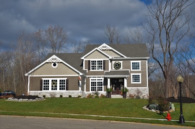 A two-story house is set against a backdrop of bare trees under a cloudy sky. The house features taupe siding with white trim and has wreaths hanging on the door and windows, suggesting a festive season. The front yard is well-kept with green grass and minimal landscaping, including some bushes and a small stone border around part of the garden. A red fire hydrant and a streetlight are visible in the foreground on the sidewalk.