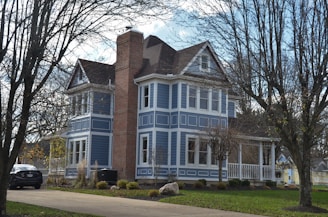 A two-story house painted in a vibrant blue color with white trim around the windows and doors. It features a prominent brick chimney and a small porch with a railing. The front yard is landscaped with a green lawn, shrubs, and trees that have shed their leaves. A car is parked in the driveway next to the house, with a cloudy sky in the background.