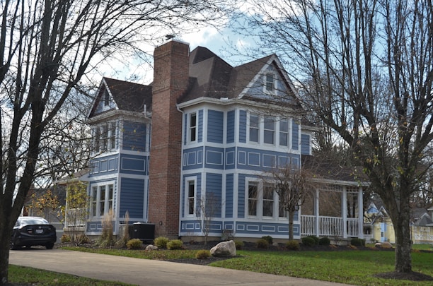 A two-story house painted in a vibrant blue color with white trim around the windows and doors. It features a prominent brick chimney and a small porch with a railing. The front yard is landscaped with a green lawn, shrubs, and trees that have shed their leaves. A car is parked in the driveway next to the house, with a cloudy sky in the background.