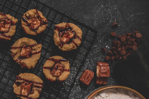 Freshly baked cookies with chocolate pieces and drizzle sitting on a cooling rack. A small pile of chocolate chips is nearby, alongside two broken pieces of chocolate bar. The dark background complements the warm tones of the cookies and chocolate.