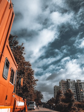 An urban street scene features large orange construction vehicles parked along a road with a backdrop of tall buildings. The sky is filled with dramatic and somewhat overcast clouds. Trees line the street, adding greenery to the scene, while pedestrians and other smaller vehicles are visible in the distance.