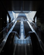 An escalator ascends towards a large, domed building seen through a wide opening at the top. The scene is framed by the dark interior of the structure housing the escalator, enhancing the contrast with the bright exterior.
