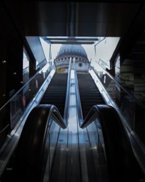 An escalator ascends towards a large, domed building seen through a wide opening at the top. The scene is framed by the dark interior of the structure housing the escalator, enhancing the contrast with the bright exterior.