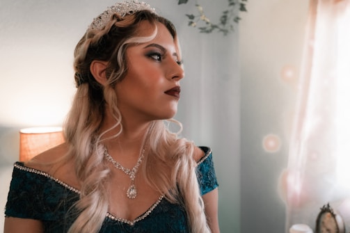 A woman adorned in a sparkling tiara and elegant jewelry gazes thoughtfully, wearing a teal off-the-shoulder dress. Her long, wavy blonde hair frames her face. The background suggests a softly lit room with a lamp and some greenery.