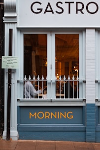 A storefront of a restaurant or café named 'Gastro' with a window showing the interior where two people can be seen sitting. The exterior is painted in a light blue with the word 'MORNING' prominently displayed.