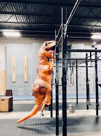 A person in a dinosaur costume is hanging from a pull-up bar in a gym environment. The gym features include climbing walls, hanging rings, and gym mats. The ceiling is industrial with metal beams, and the lighting is bright.