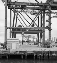 A large industrial crane structure dominates the image, with intricate metal beams and support systems visible. In the background, a city skyline is partially visible, including a few skyscrapers. A dock with concrete barriers and water is in the foreground.