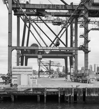 A large industrial crane structure dominates the image, with intricate metal beams and support systems visible. In the background, a city skyline is partially visible, including a few skyscrapers. A dock with concrete barriers and water is in the foreground.