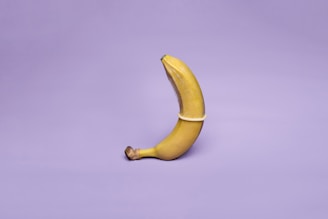 A single banana stands upright against a plain lavender background, covered by a translucent condom, illustrating a symbolic representation.