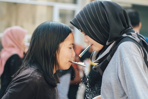 Two people are engaging in a playful activity involving straws and crackers. They are facing each other closely, each holding a cracker with a straw. The focus is on their interaction and concentration. In the background, other people are visible, indicating a social setting.