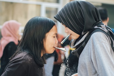 Two people are engaging in a playful activity involving straws and crackers. They are facing each other closely, each holding a cracker with a straw. The focus is on their interaction and concentration. In the background, other people are visible, indicating a social setting.