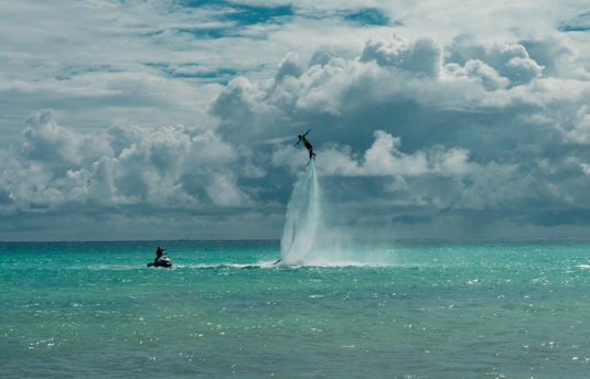 A person is flying high above the ocean using a water jetpack, propelled by powerful jets of water. The sky is filled with dramatic, thick clouds, adding a sense of adventure and intensity to the scene. Nearby, another person is riding a jet ski on the turquoise water.