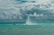 A person is flying high above the ocean using a water jetpack, propelled by powerful jets of water. The sky is filled with dramatic, thick clouds, adding a sense of adventure and intensity to the scene. Nearby, another person is riding a jet ski on the turquoise water.