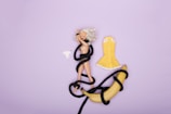 A blonde doll is wrapped in a black rope, lying on a light purple background. The doll is unclothed, and separate pieces of clothing, including a pair of panties and a yellow skirt, are placed nearby. A banana is also entwined with the same rope.