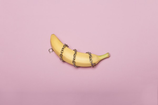 A banana is placed on a pink background. It is wrapped with a metal chain that loops around its middle, creating a contrast between the natural fruit and the cold metal.