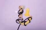 A blonde doll is lying on a purple surface, entangled with a black rope. Beside the doll, there is a yellow dress, a pair of white panties, and a banana also partly wrapped in the black rope.