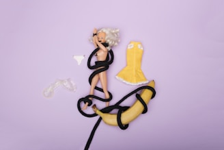 A blonde doll is lying on a purple surface, entangled with a black rope. Beside the doll, there is a yellow dress, a pair of white panties, and a banana also partly wrapped in the black rope.