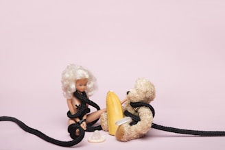 A doll with white hair and light skin is seated on the floor, entangled in a black rope. Beside the doll is a plush teddy bear, also wrapped in the same black rope. In between them lies a yellow object resembling a surfboard or some kind of paddling equipment, and in front of the doll is a white plastic object that looks like a baby bottle nipple or a pacifier. The background is a plain light pink color.