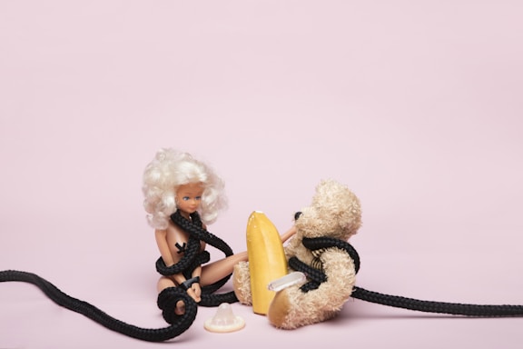A doll with white hair and light skin is seated on the floor, entangled in a black rope. Beside the doll is a plush teddy bear, also wrapped in the same black rope. In between them lies a yellow object resembling a surfboard or some kind of paddling equipment, and in front of the doll is a white plastic object that looks like a baby bottle nipple or a pacifier. The background is a plain light pink color.