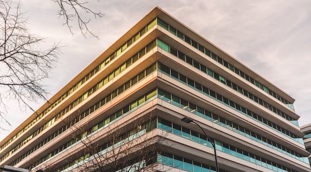 A multi-story office building with a modern architectural design. The structure features large glass windows and horizontal lines. Surrounding the building are bare tree branches and a streetlight, suggesting a cityscape environment.