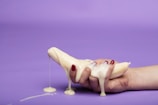 A hand with red-painted fingernails holds a peeled banana covered in a thick, creamy substance against a solid purple background. The cream is dripping down the banana and fingers, creating a messy and somewhat provocative composition.