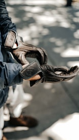 A person wearing dark brown gloves, possibly made of leather, with a textured pattern. The person is dressed in a dark jacket, and the background features a bokeh effect with dappled sunlight on the pavement.
