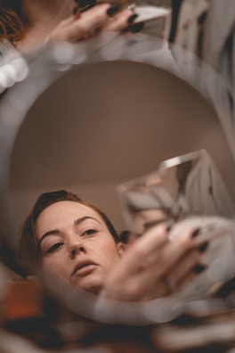 A person is reflected in a round mirror, gazing pensively while holding a glass object. The focus is soft, creating an introspective mood. Black nail polish is noticeable on their fingers.