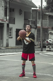 A man is standing on a residential street basketball court, holding a basketball in his hands as he prepares to take a shot. He is wearing a black sports jersey, red shorts, and red sports shoes. Buildings and a parked car can be seen in the background, with the mood suggesting a casual neighborhood setting.