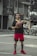A man stands on an outdoor basketball court holding a basketball. He is dressed in athletic clothing including a black shirt, red shorts, and red athletic shoes. The background features residential buildings and a partially visible car.