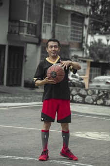 A man stands on an outdoor basketball court holding a basketball. He is dressed in athletic clothing including a black shirt, red shorts, and red athletic shoes. The background features residential buildings and a partially visible car.