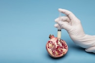 A gloved hand holds a syringe filled with a creamy white substance, injecting it into a pomegranate half resting on a light blue background. The pomegranate is open, revealing its seeds and vibrant red interior.