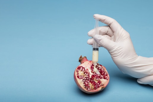 A gloved hand holds a syringe filled with a creamy white substance, injecting it into a pomegranate half resting on a light blue background. The pomegranate is open, revealing its seeds and vibrant red interior.