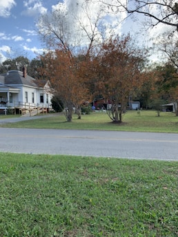 A suburban scene with a house on the left featuring a porch and wooden accessibility ramp. The yard has well-maintained grass and several trees with autumnal foliage. The sky is partly cloudy, and there are some outdoor items near the trees, including a small shed.