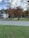 A suburban scene with a house on the left featuring a porch and wooden accessibility ramp. The yard has well-maintained grass and several trees with autumnal foliage. The sky is partly cloudy, and there are some outdoor items near the trees, including a small shed.