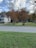 A suburban scene with a house on the left featuring a porch and wooden accessibility ramp. The yard has well-maintained grass and several trees with autumnal foliage. The sky is partly cloudy, and there are some outdoor items near the trees, including a small shed.