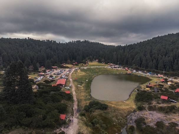 A scenic landscape with a small body of water surrounded by lush green forest. Numerous small buildings with red roofs are scattered around the area, along with a visible dirt path leading through the vegetation. Several vehicles are parked on the grassland, suggesting a camping or recreational area. The sky above is overcast with dark clouds, adding a dramatic effect to the scene.