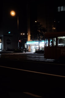 A dimly lit street scene at night featuring a brightly illuminated convenience store, with its sign reading 'FamilyMart'. A quiet street crossing in the foreground with faint outlines of a bus or large vehicle to the right and some pedestrians visible. Streetlights cast a warm glow over the area, while other buildings remain in shadow.