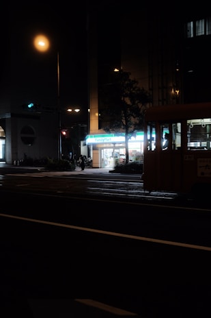 A dimly lit street scene at night featuring a brightly illuminated convenience store, with its sign reading 'FamilyMart'. A quiet street crossing in the foreground with faint outlines of a bus or large vehicle to the right and some pedestrians visible. Streetlights cast a warm glow over the area, while other buildings remain in shadow.