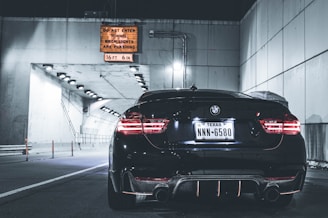 A black BMW car with Texas license plates is parked near the entrance of a tunnel. The tunnel has bright lights along the ceiling, casting a cool illumination. A warning sign above the entrance specifies height restrictions and instructions for when the tunnel is closed.