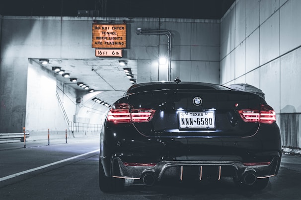 A black BMW car with Texas license plates is parked near the entrance of a tunnel. The tunnel has bright lights along the ceiling, casting a cool illumination. A warning sign above the entrance specifies height restrictions and instructions for when the tunnel is closed.