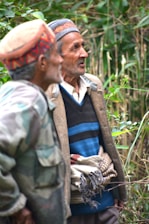Two elderly men stand side by side, wearing traditional hats and layered clothing. One wears a patterned cardigan over a striped sweater, while the other has a camouflage jacket. The background features dense greenery, and both men are focused on something out of frame, possibly engaged in conversation.