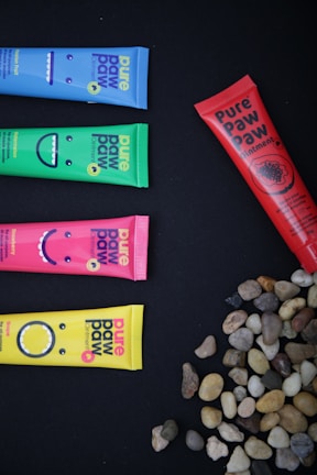 Colorful tubes of Pure Paw Paw ointment are arranged on a black background. There are four horizontal tubes in blue, green, pink, and yellow, each with smiling face designs. A red tube is positioned vertically alongside scattered small stones in a variety of natural shades.