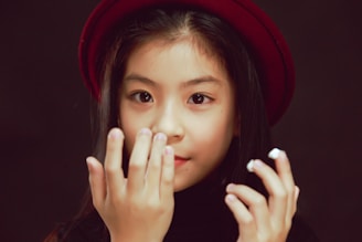 A young girl is wearing a dark outfit and a maroon hat, with long black hair and an expression that suggests curiosity or contemplation. Her hands are raised near her face.