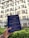 A hand holding a blue coffee packaging labeled with 'Fisher Coffee' in front of a building with multiple windows and air conditioning units. The packaging contains text about coffee roasting. The background shows greenery and part of a sunny sky.