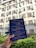 A hand holding a blue coffee packaging labeled with 'Fisher Coffee' in front of a building with multiple windows and air conditioning units. The packaging contains text about coffee roasting. The background shows greenery and part of a sunny sky.