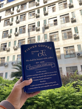 A hand holding a blue coffee packaging labeled with 'Fisher Coffee' in front of a building with multiple windows and air conditioning units. The packaging contains text about coffee roasting. The background shows greenery and part of a sunny sky.