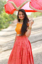 A relaxed model wearing a bright orange blouse standing in a sunny outdoor market.